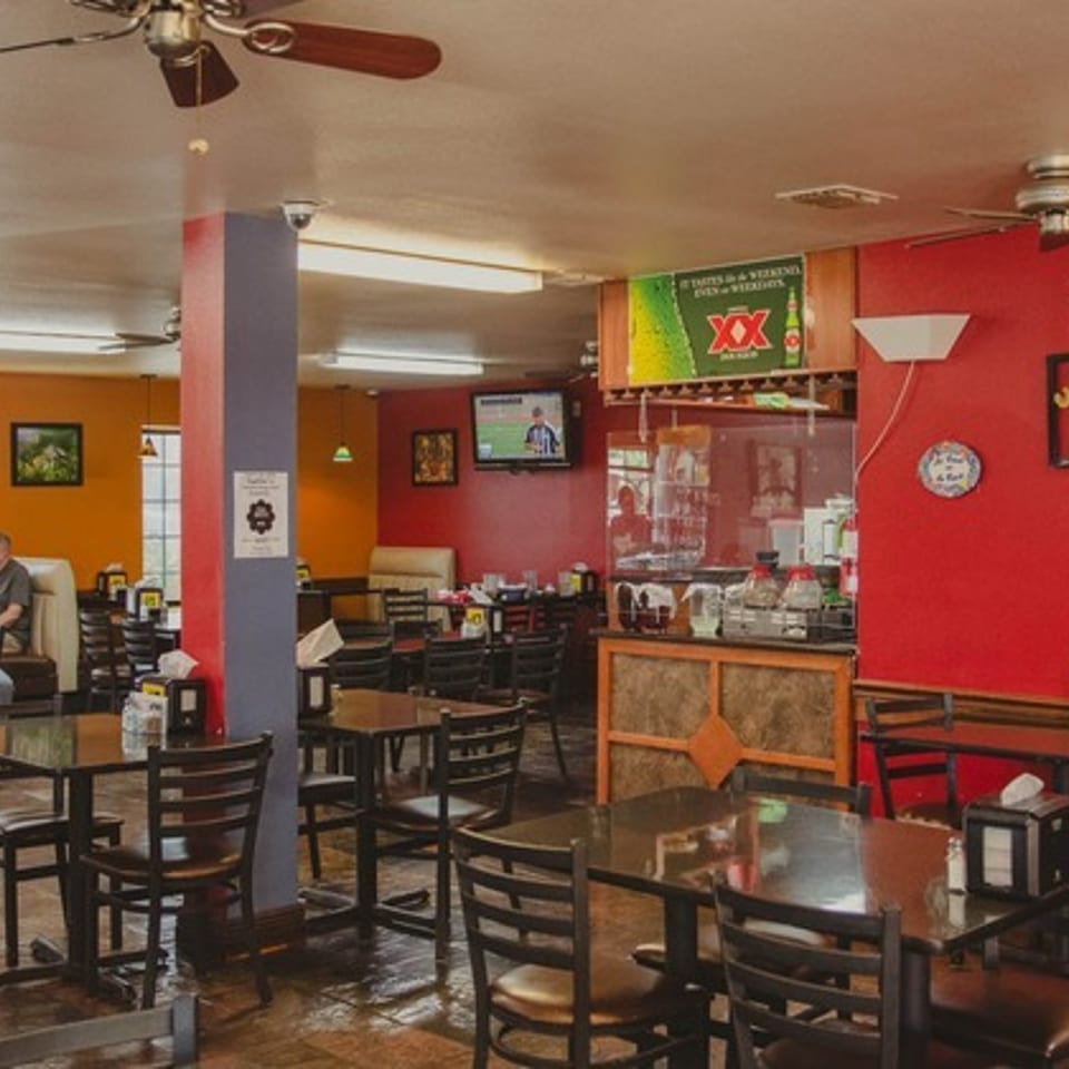 Colorful casual Mexican restaurant dining room with black tables, seated diners, ceiling fans, and a prominent Dos Equis beer sign.