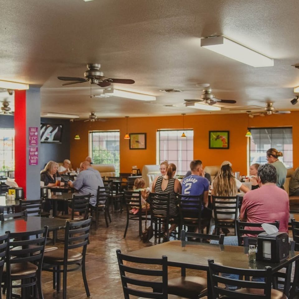 Busy casual restaurant dining room with orange walls, ceiling fans, wooden tables, and patrons seated near sunlit windows.